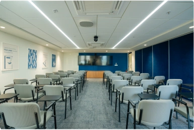 Empty conference room with rows of gray chairs facing a large screen and blue walls at Manubhai & Shah LLP office.