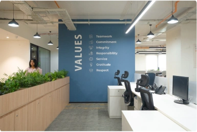 Modern Manubhai & Shah LLP office with desks, chairs, a blue wall listing core values, and plants in front of a woman.