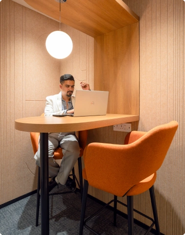 Man in a white blazer working on a laptop in a small wooden workspace with orange chairs at Manubhai & Shah LLP office.