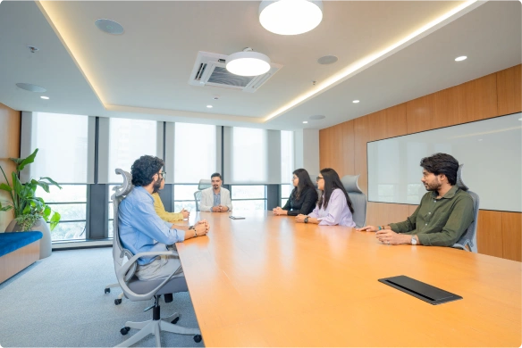 Five people engaged in a meeting around a large wooden conference table in a modern Manubhai & Shah LLP office.