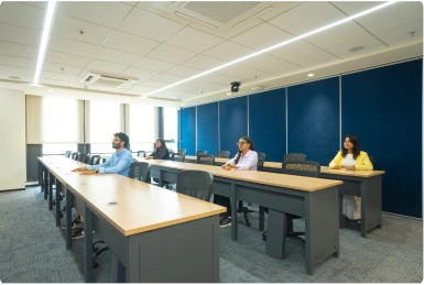 Three people seated at desks in a spaced, modern classroom with large windows and blue walls at Manubhai & Shah LLP office.