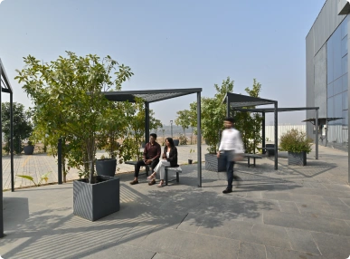 Two people sitting under black pergolas with plants nearby and one person walking on a paved outdoor area at Deakin University.