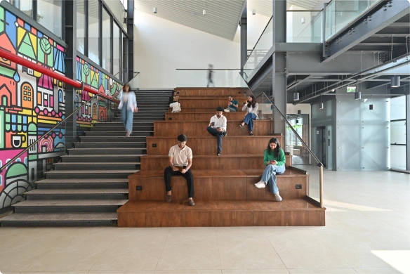People sitting and walking on tiered wooden seating next to colorful mural in a modern indoor space at Deakin University.