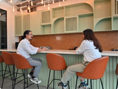 Two people sitting on orange bar stools, talking in a modern kitchen with green cabinets and hanging lights at Deakin University.