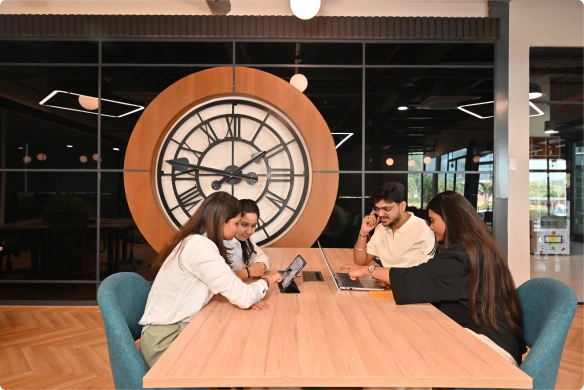 Four people working together at a table with laptops and a large clock on the wall behind them at Deakin University.