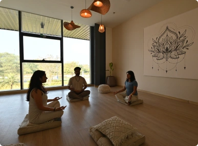 Three people meditating on cushions in a spacious room with large windows and wall art at Deakin University.