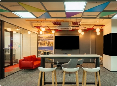 Walter P Moore modern office lounge with high table, stools, gray and red chairs, ceiling lights, and a TV on the wall.