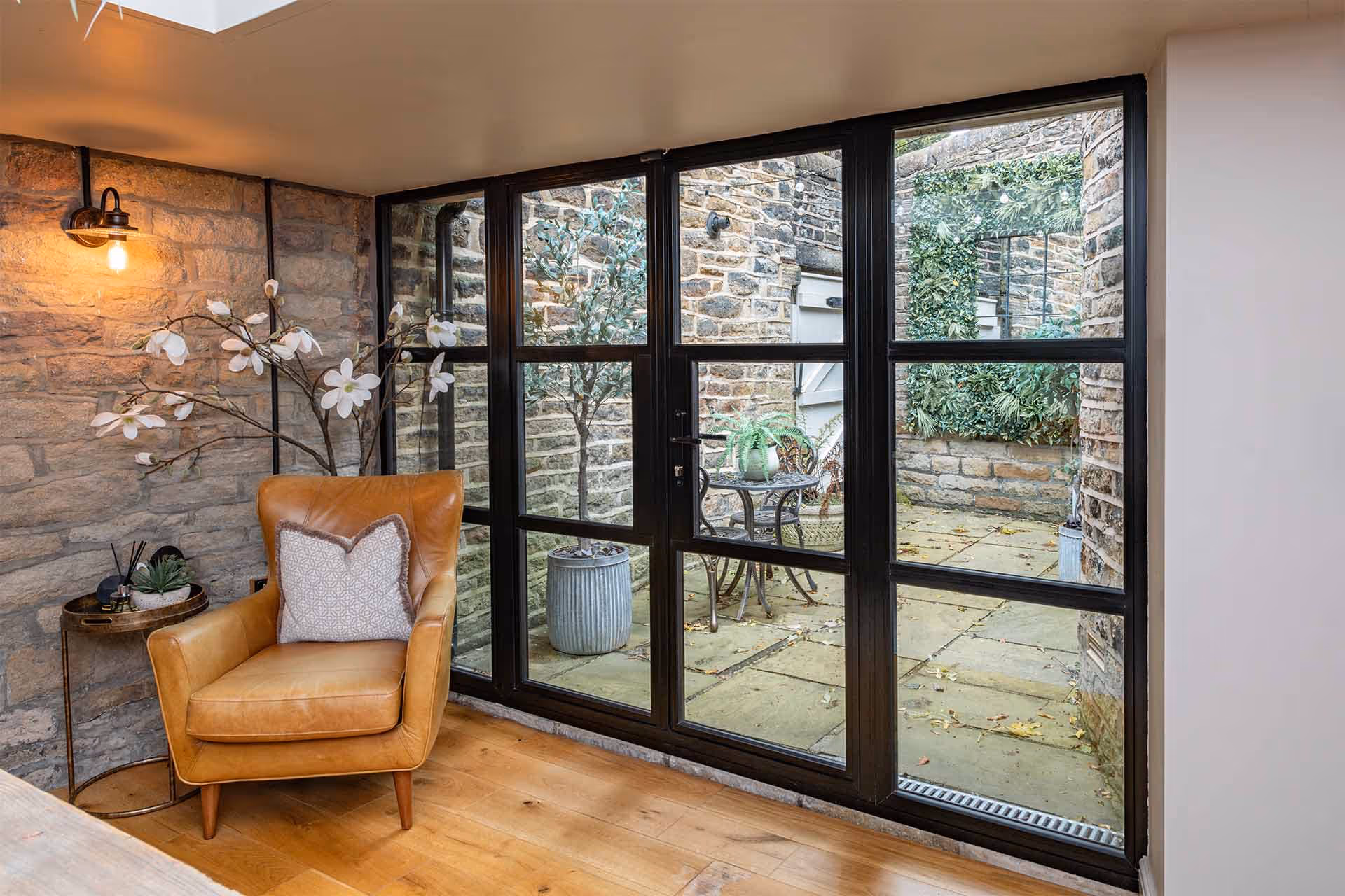 Black heritage-style aluminium doors by Decorio opening from a cosy living space with stone walls onto a traditional courtyard patio