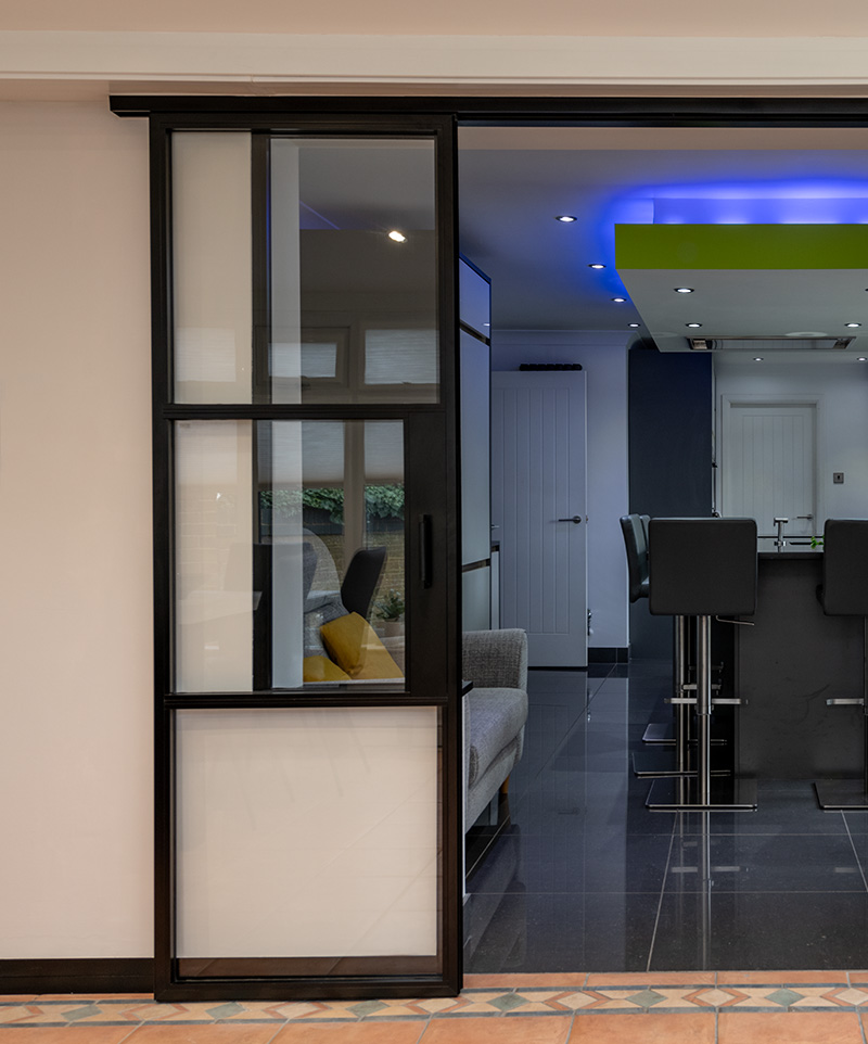 Modern black-framed sliding door opening into a kitchen with dark flooring, bar stools, and blue ceiling lights.
