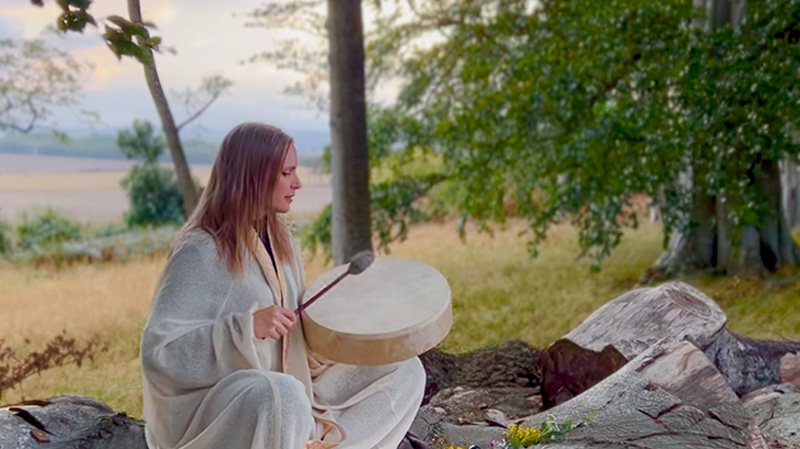 Shamanic practitioner in white cloak playing a drum in the forest, honoring Norse traditions during the fall equinox.