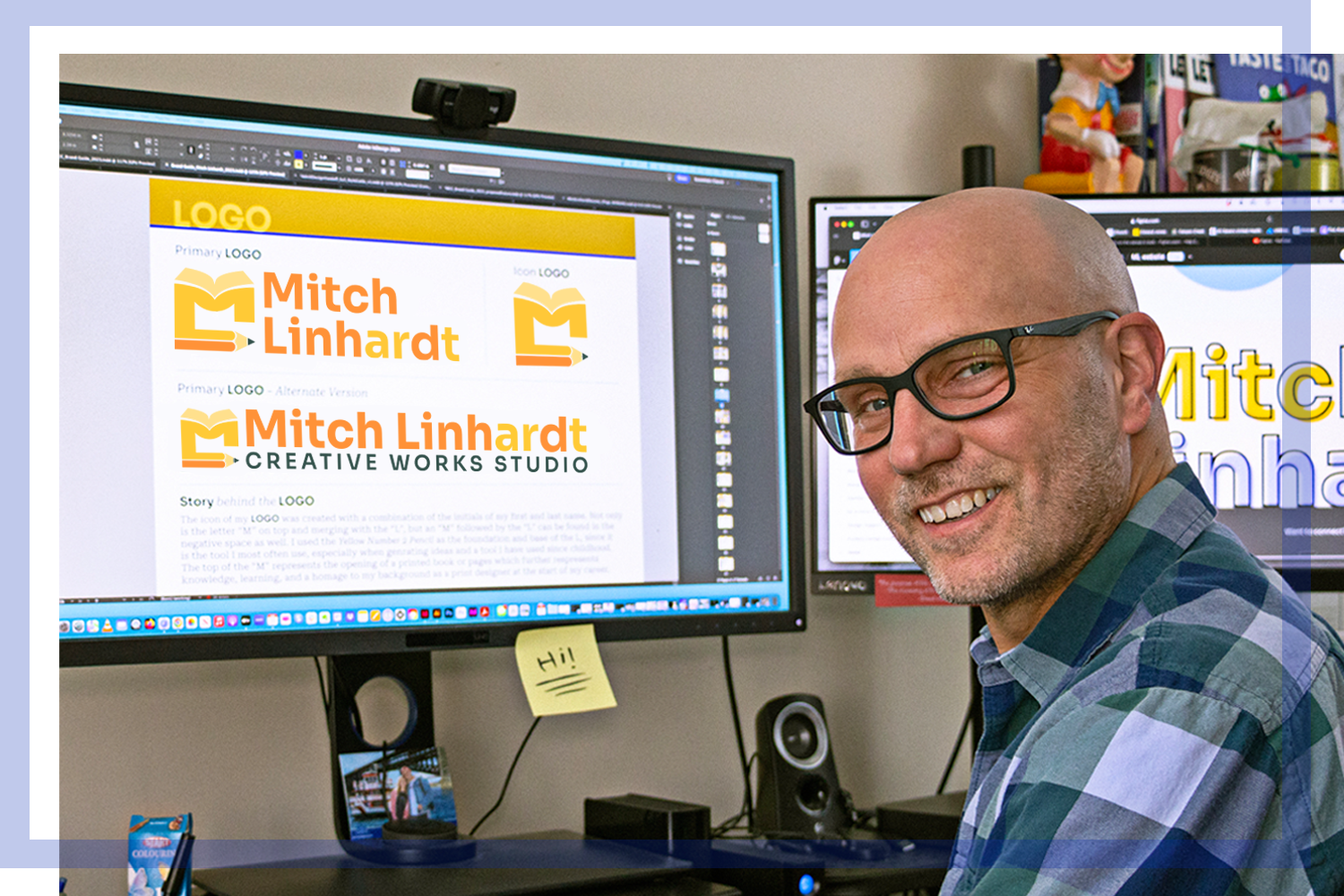Smiling man wearing glasses and a checkered shirt sitting at a desk with two monitors displaying logo designs for 'Mitch Linhardt Creative Works Studio' and other personal workspace items.