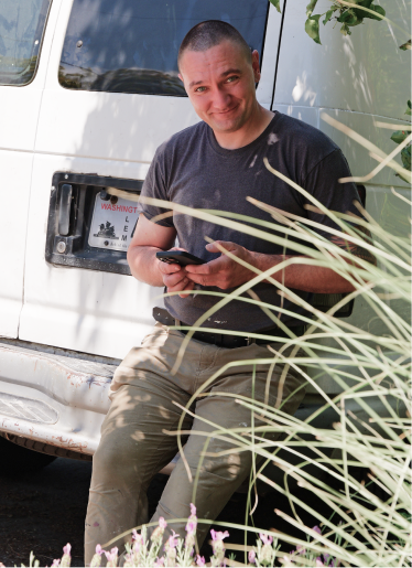 Man smiling while leaning against a white work van, holding a phone. Plants partially obscure the foreground, creating a candid and relaxed scene.