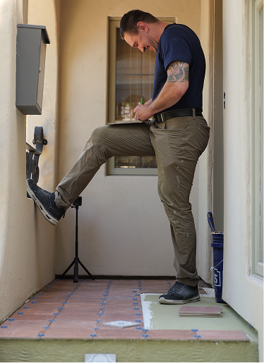 Man standing on a tiled front porch, reviewing notes on a clipboard with one foot propped on a railing. Early stage of tile layout visible beneath him.