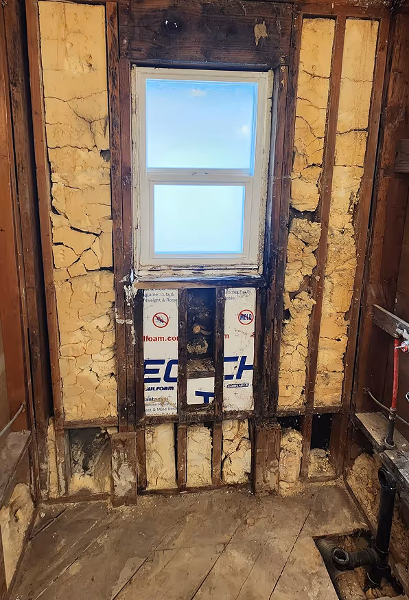 Shower walls taken down to the studs during a bathroom demolition, exposing framing and insulation around a window.