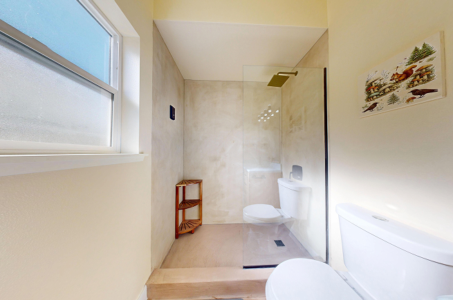 Completed walk-in microcement shower with frameless glass panel and brass rain showerhead in West Seattle rental bathroom.