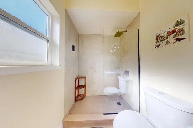 Completed walk-in microcement shower with frameless glass panel and brass rain showerhead in West Seattle rental bathroom.