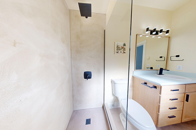Microcement shower with matte black fixtures and frameless glass panel next to updated light oak vanity in West Seattle bathroom.
