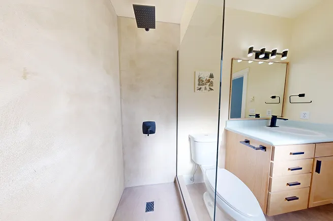 Microcement shower with matte black fixtures and frameless glass panel next to updated light oak vanity in West Seattle bathroom.