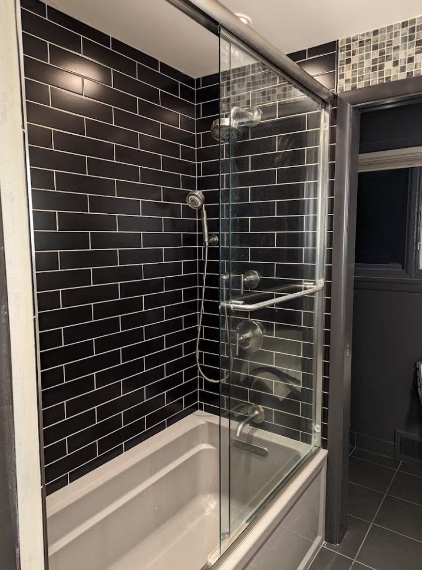 Completed tub and shower in dark subway tile with a glass panel and mosaic accent strip.