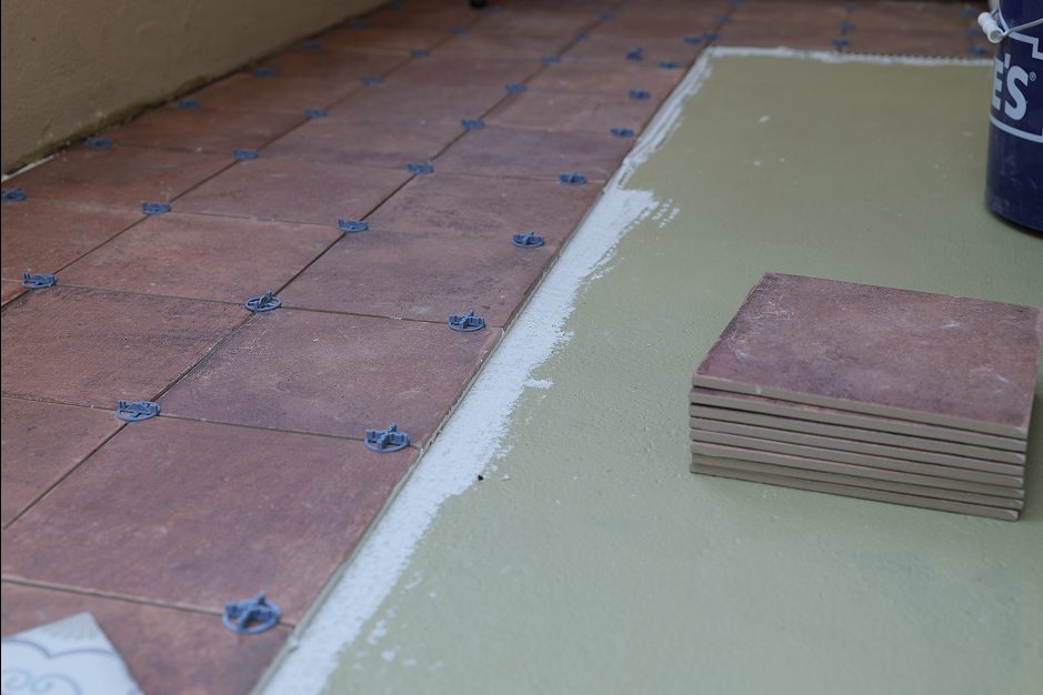 In-progress view of terracotta tiles being laid on a green-painted surface using blue leveling spacers. A small stack of matching tiles sits nearby next to a bucket.