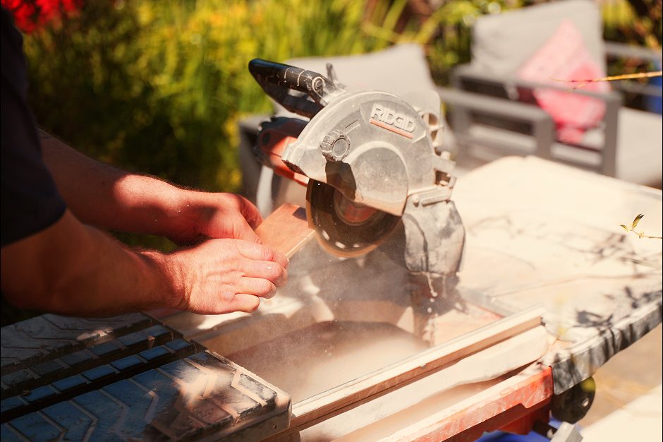 Man using a RIDGID wet tile saw outdoors to make precise cuts on a tile. The saw is positioned on an orange workbench surrounded by greenery and sunlight.