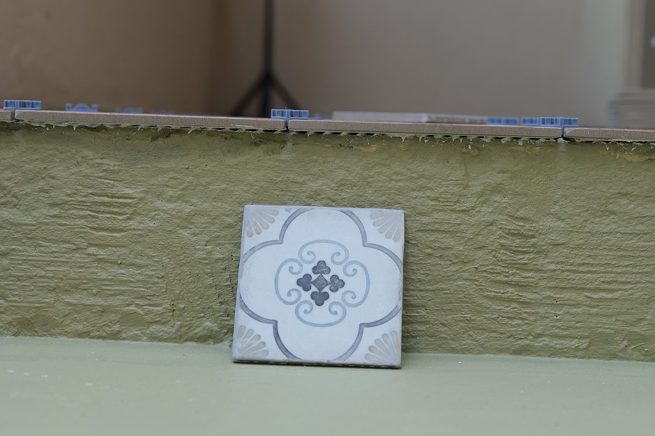 Single patterned porcelain tile leaning against a green-painted stair riser before installation, with partially laid tiles and leveling spacers visible above.