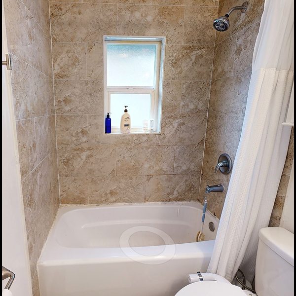 Before renovation: beige-tiled tub-shower with frosted window, chrome fixtures, white curtain, and a few toiletry bottles on the sill.