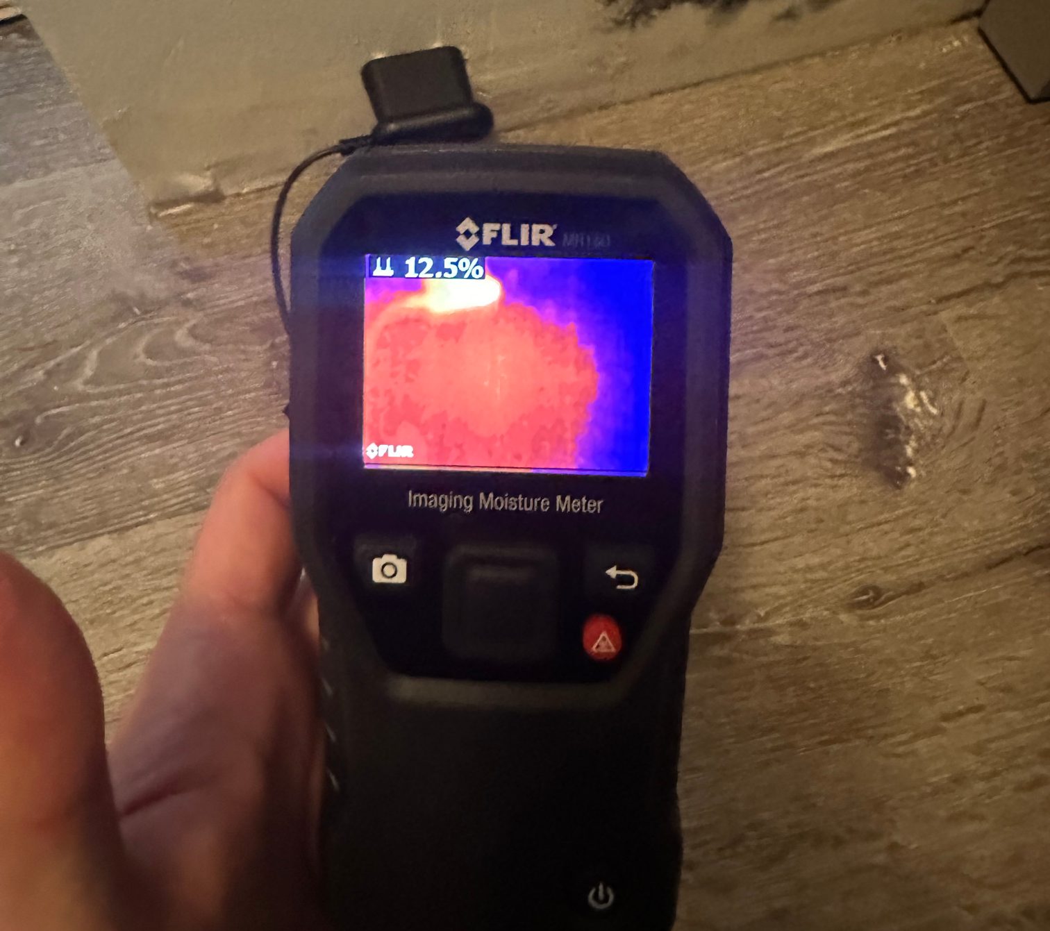 Handheld thermal imaging moisture meter reading 12.5 percent on a wood floor beside a shower.