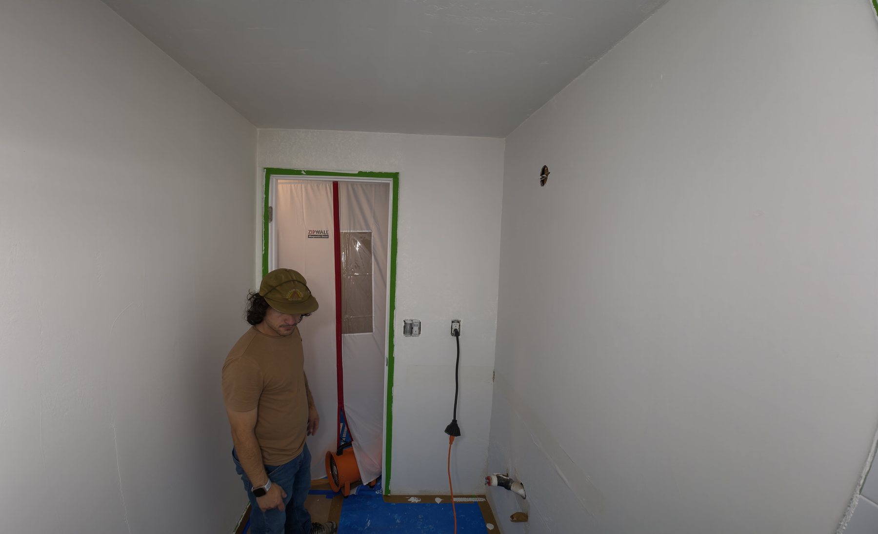 A West Seattle bathroom mid-renovation: drywall up and unfinished, a dust barrier taped across the doorway, floor protection down, plumbing stubbed out at the floor, and a dedicated outlet on the wall with a cord running from it.