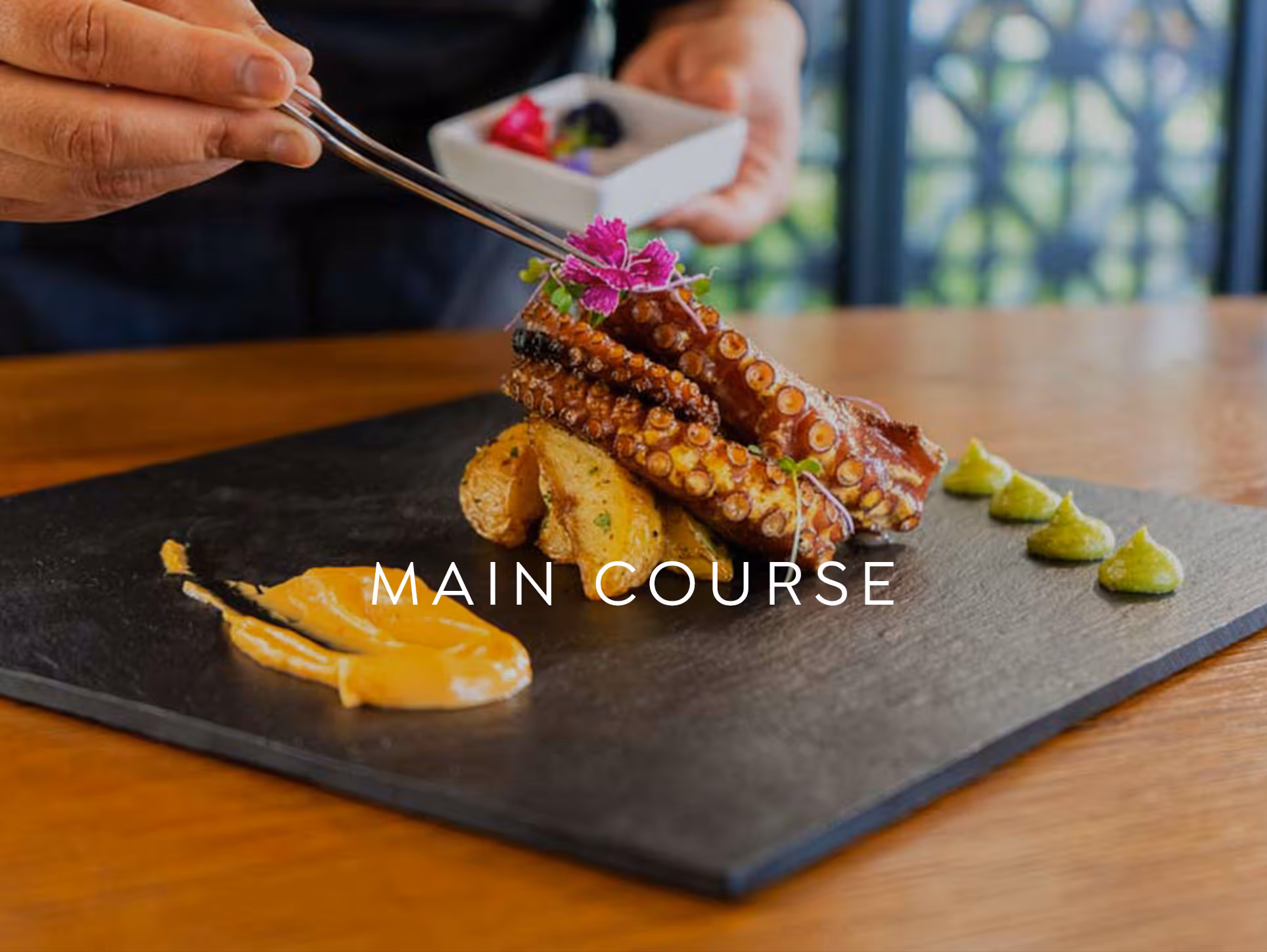 Grilled octopus tentacles on roasted potatoes with sauce, garnished with edible flowers and green herb dots on a black slate plate.