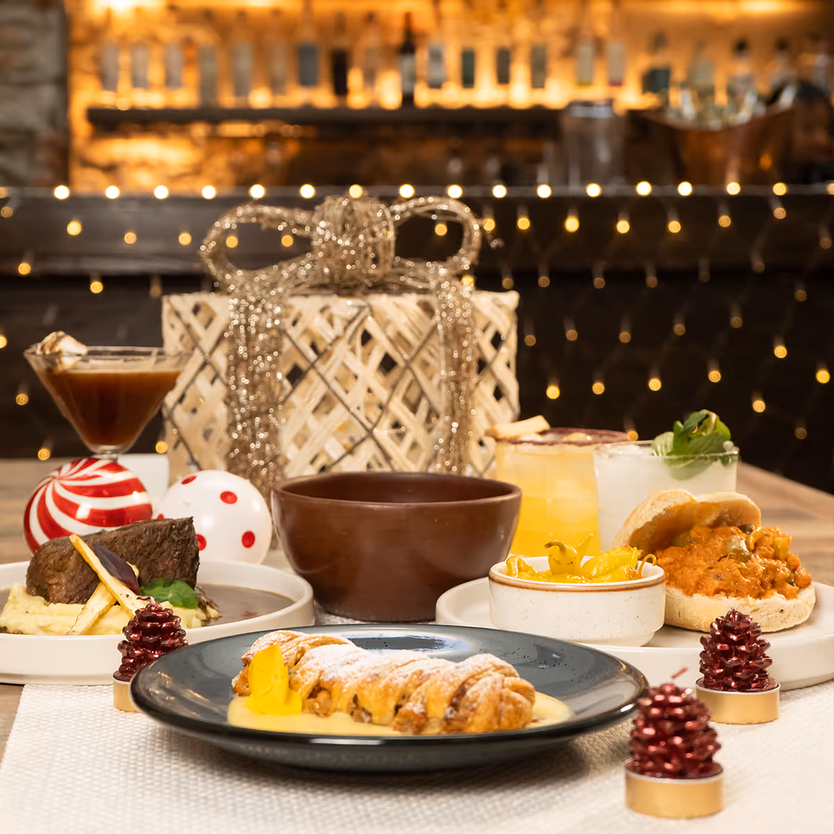 Festive table setting with plated gourmet dishes, cocktails, decorative gift box, and holiday-themed ornaments.