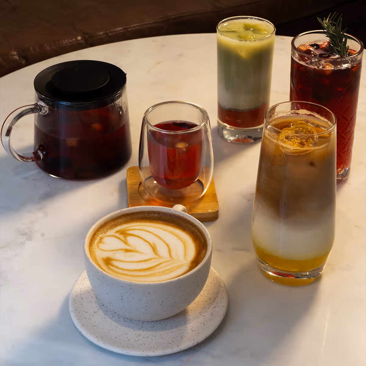 A variety of coffee and tea drinks on a marble table, including a latte with latte art, iced layered coffee, iced matcha latte, a glass of herbal tea, a glass of iced berry tea garnished with rosemary, and a teapot with tea.