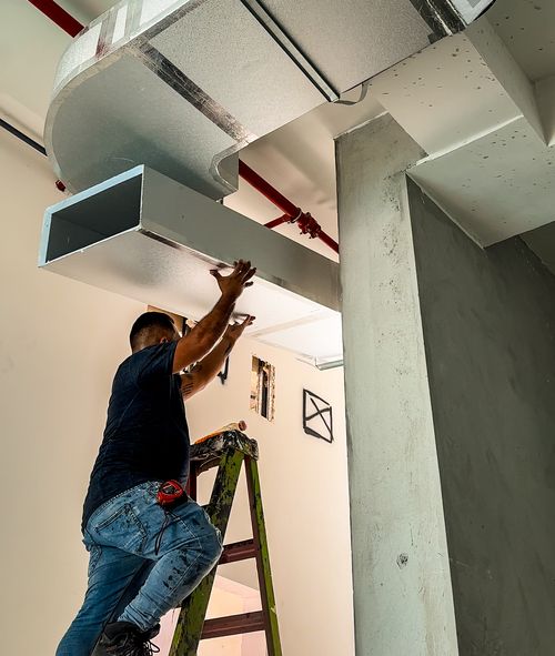 Un trabajador de SYMISA Soluciones y Mejoras Integrales S.A. está subido en una escalera instalando un conducto de ventilación metálico en el techo. Está en una posición elevada, sosteniendo el conducto con ambas manos mientras lo ajusta a la estructura superior. La imagen muestra una obra en construcción, con tuberías y conductos visibles. El trabajador lleva una camiseta oscura y jeans, y parece estar concentrado en asegurar correctamente el conducto.