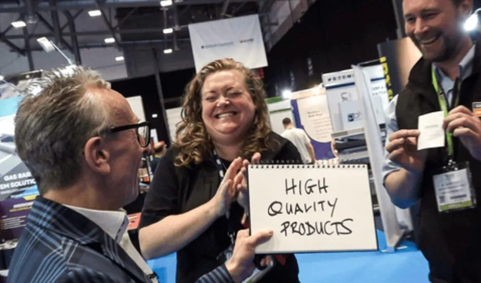 Three people at a trade show; one showing a sign reading High Quality Products and everyone smiling.