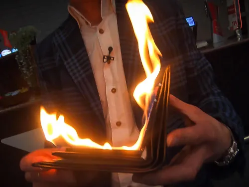Person in checkered jacket holding a burning wallet with visible flames.
