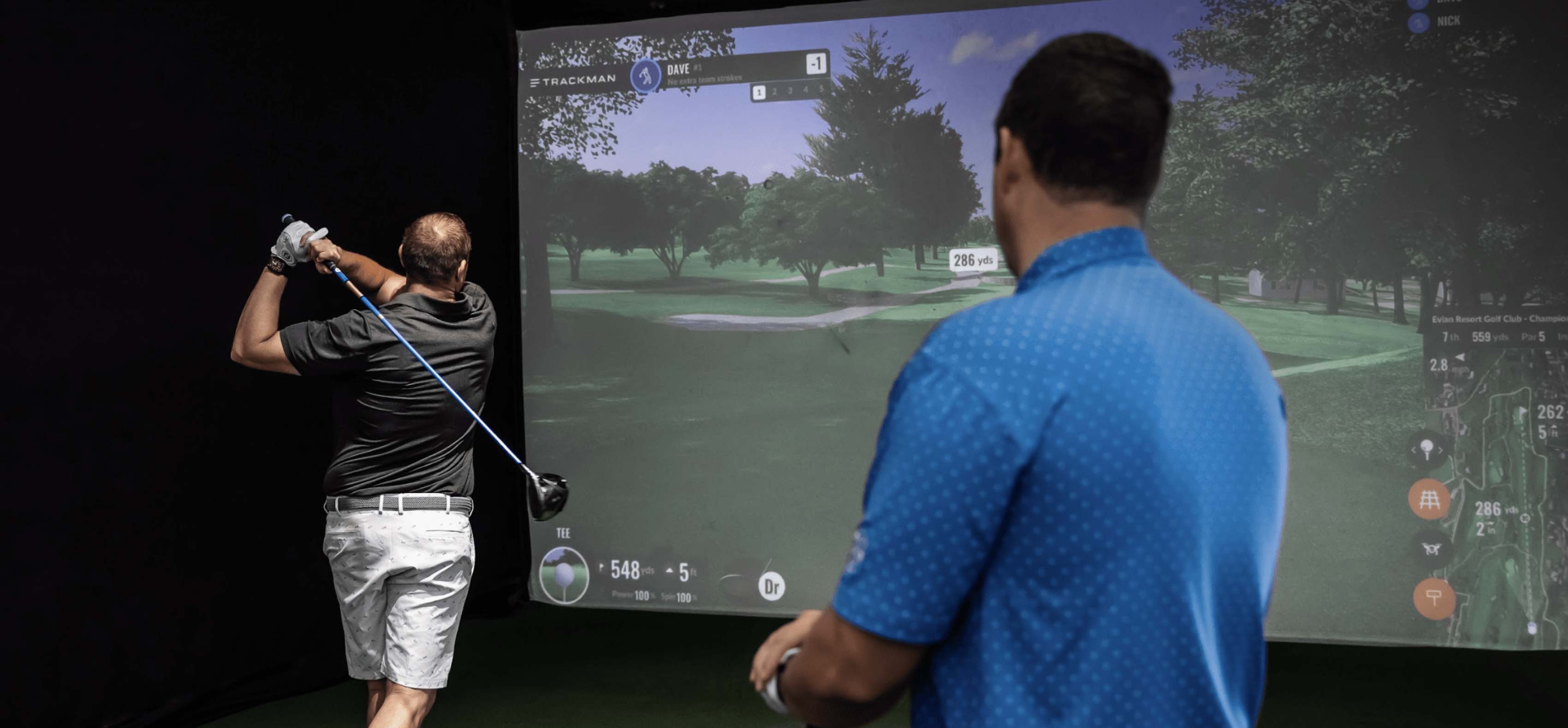 Close up view of one man finishing his golf swing towards the simulator screen while another man looks on