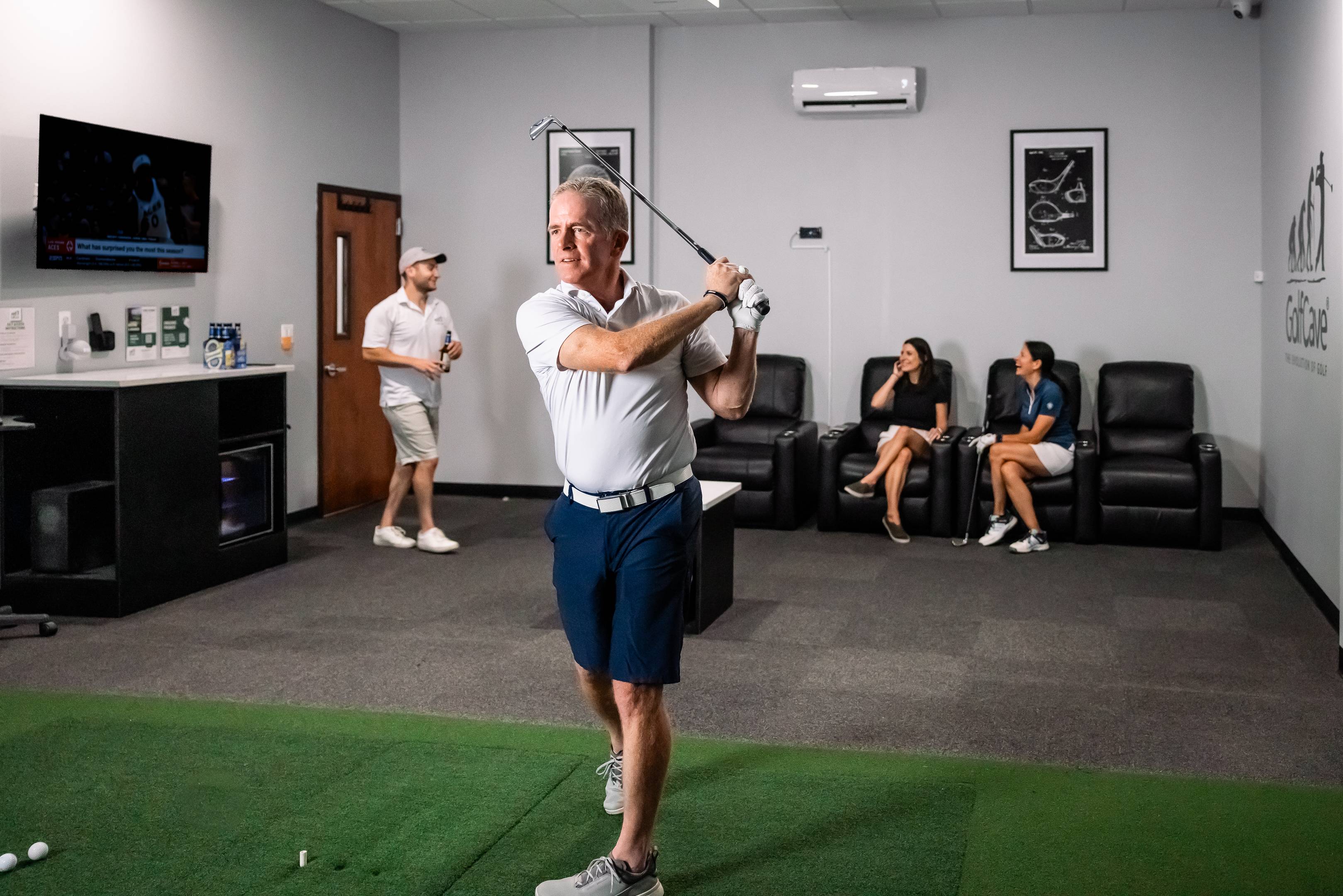Man playing inside a golf simulator room at GolfCave with a group of people talking in the background at a golf party 