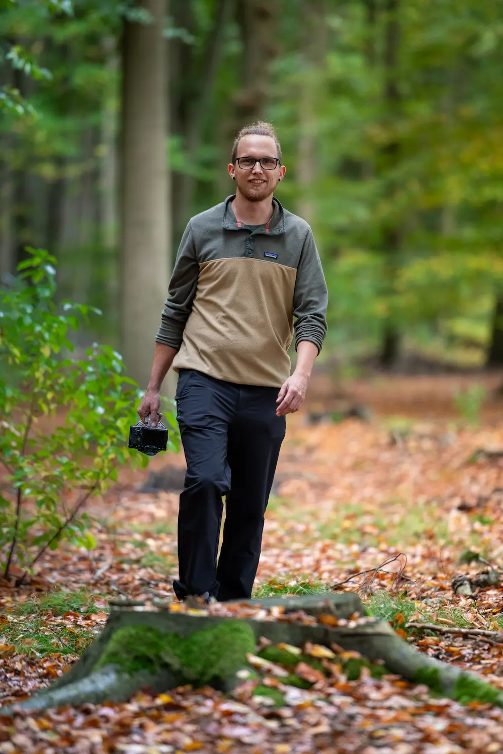 Photographer walking through a forest trail holding a camera, surrounded by autumn foliage.