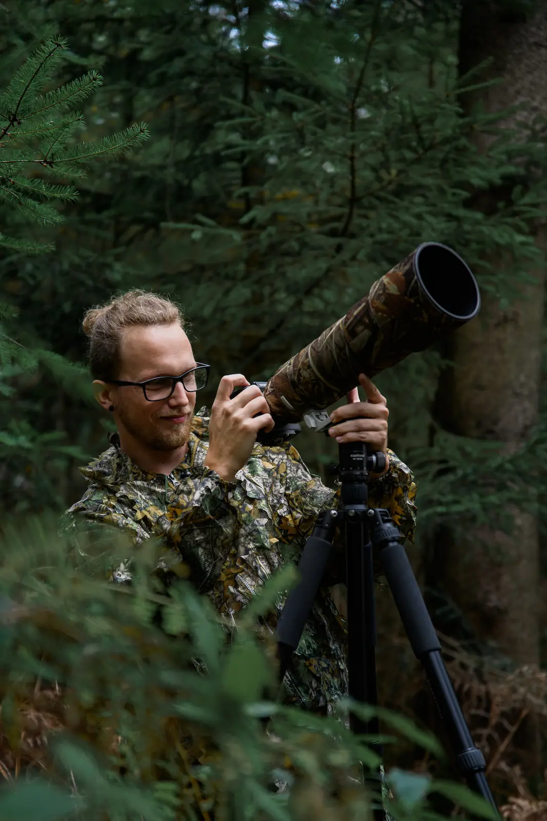 Photographer adjusting a telephoto lens on a tripod while surrounded by dense forest vegetation.