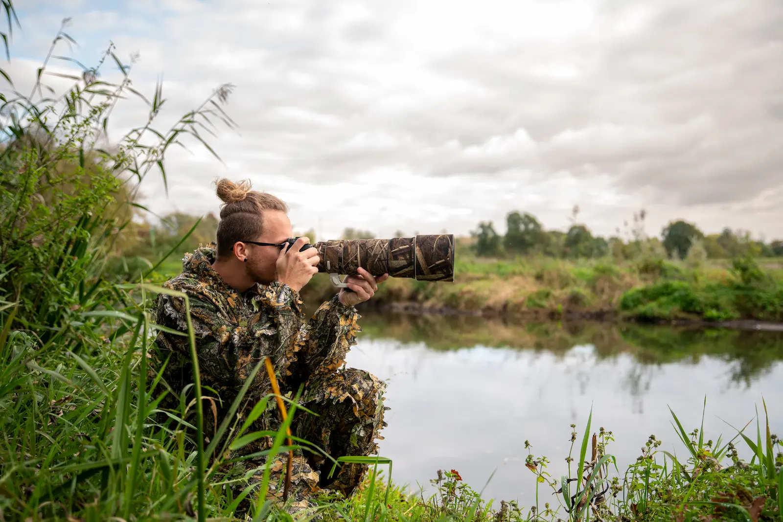 Wildlife photographer in camouflage gear crouched near a riverbank capturing nature through a telephoto lens.