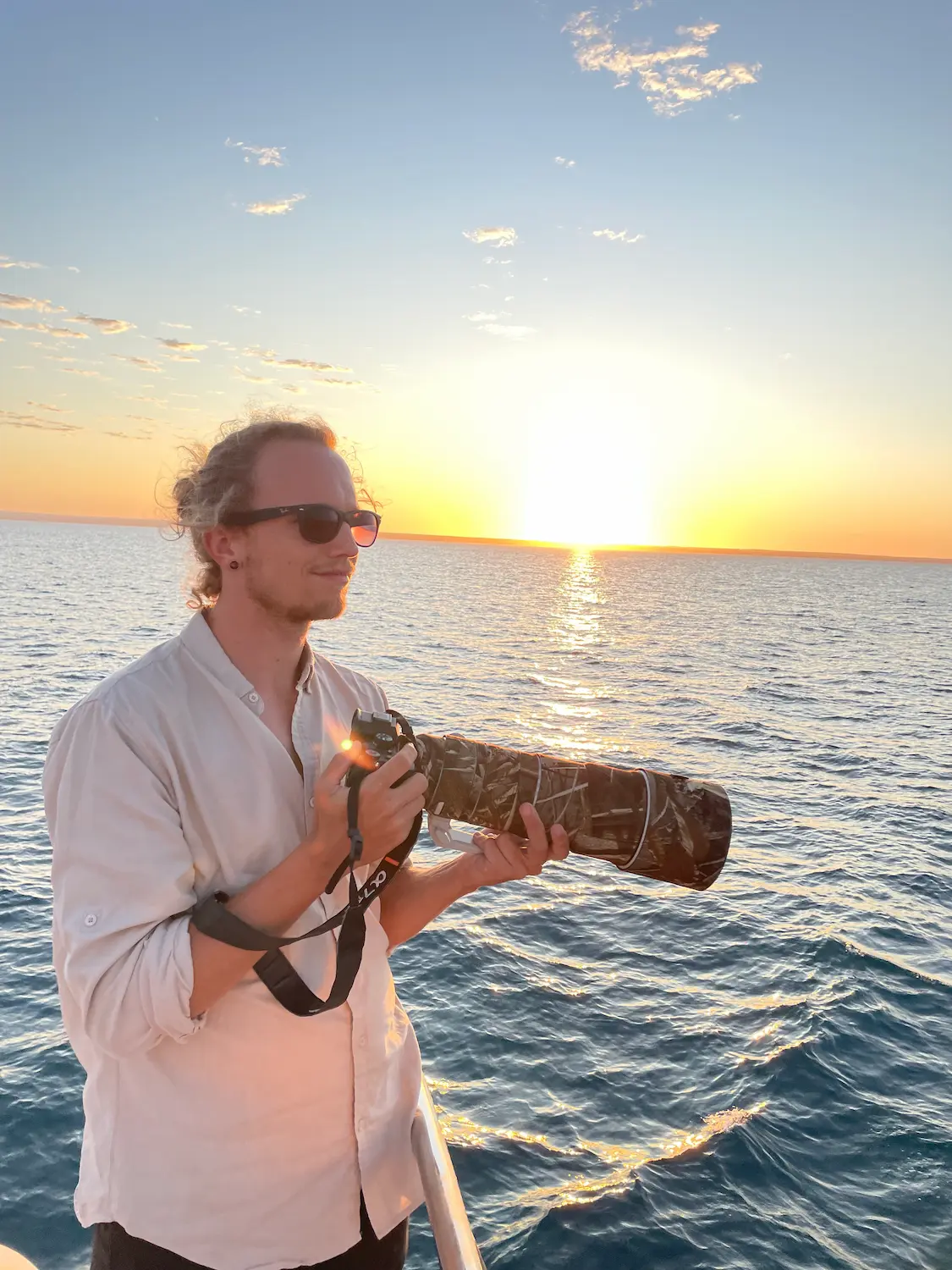Photographer standing on a boat at sunset holding a telephoto camera lens over the ocean.