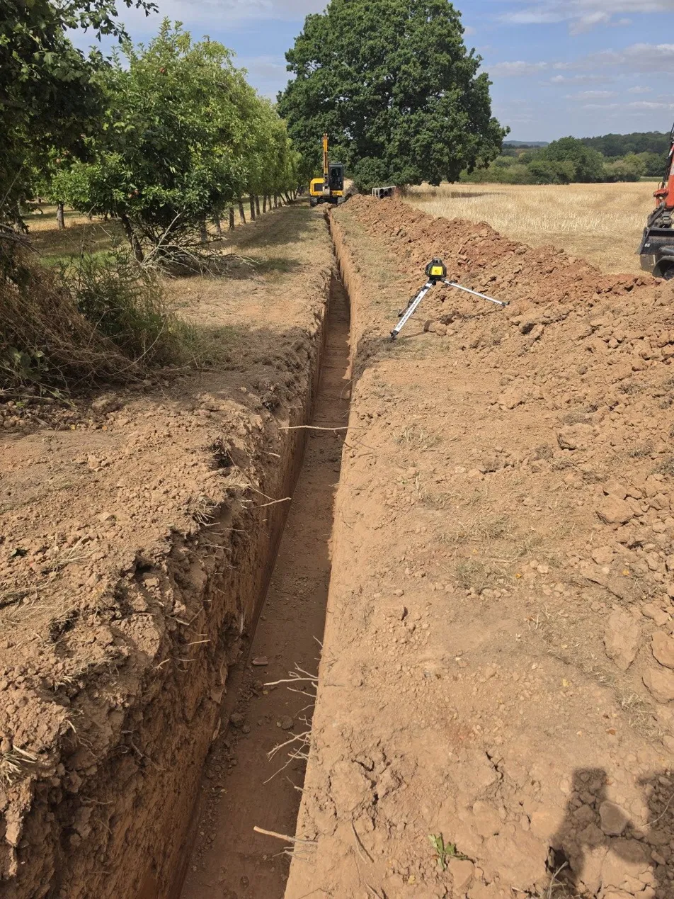Long trench excavated to level beside an orchard, with laser level set up