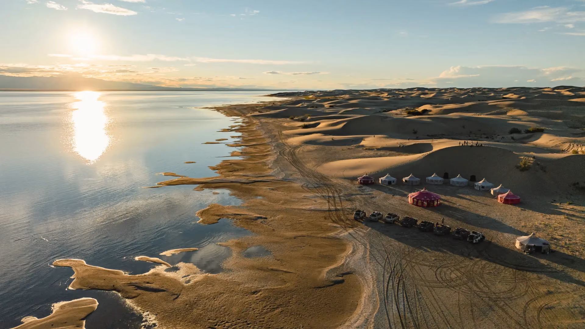 A remote overland expedition basecamp features traditional Mongolian gers nestled between rolling desert dunes and the clear waters of Durgun Lake.