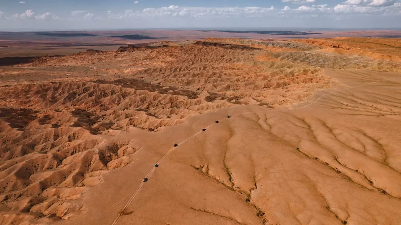 Aerial view of a vehicle convoy crossing sandy desert terrain beside eroded rock formations.