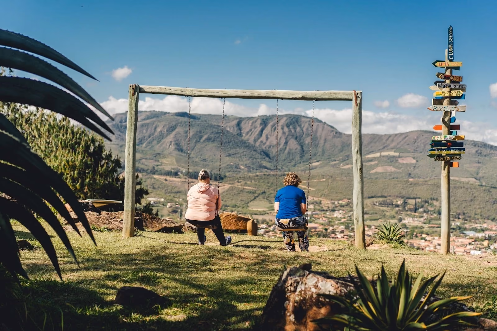 Two women sitting on a rustic wooden swing set overlooking a scenic mountain valley and a small town in Samaipata, Bolivia, with a colorful multi-directional distance signpost standing nearby on a sunny day.