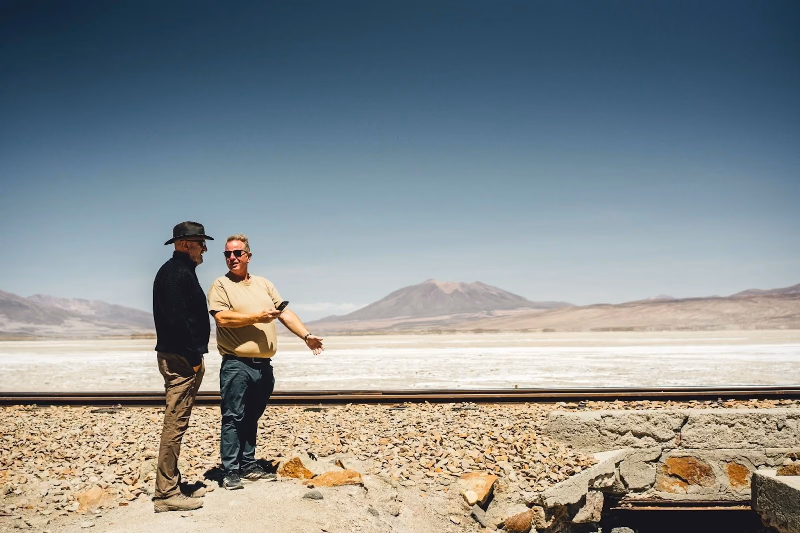 Two travellers beside a railway line overlooking an Atacama Desert salt flat.