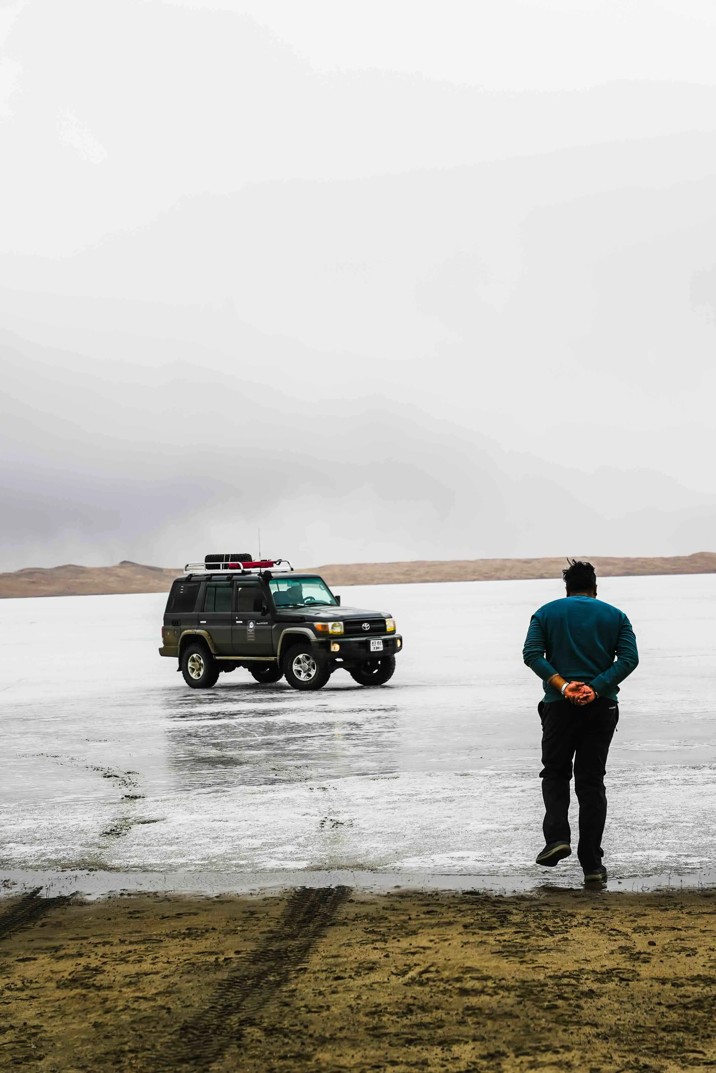 A man standing on the shore of a frozen lake facing a four-wheel-drive vehicle on the ice.