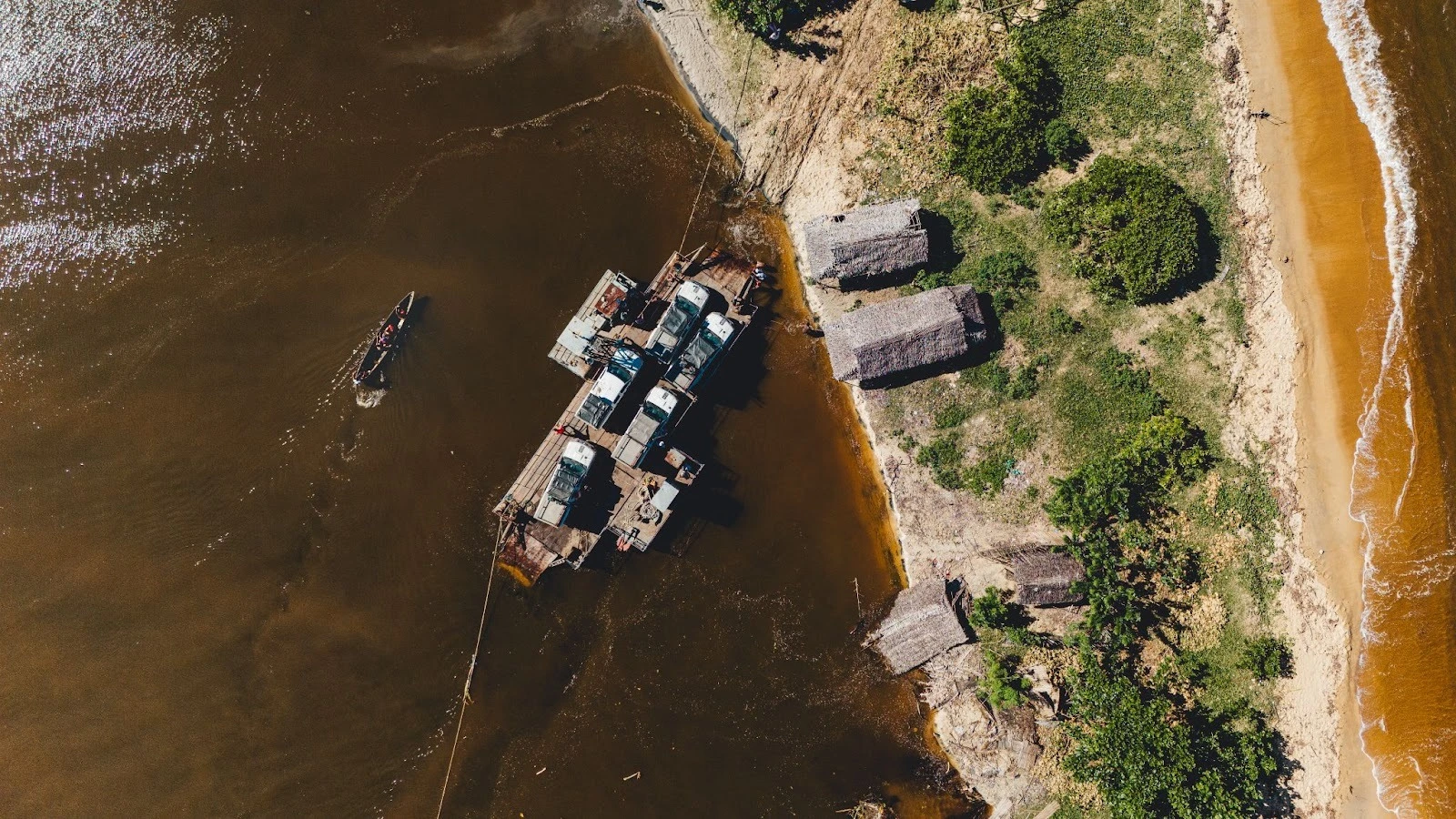 An aerial top-down view of a rustic cable ferry carrying multiple 4x4 trucks across a brown river to a sandy bank with thatched-roof huts along the RN5 route in Madagascar.