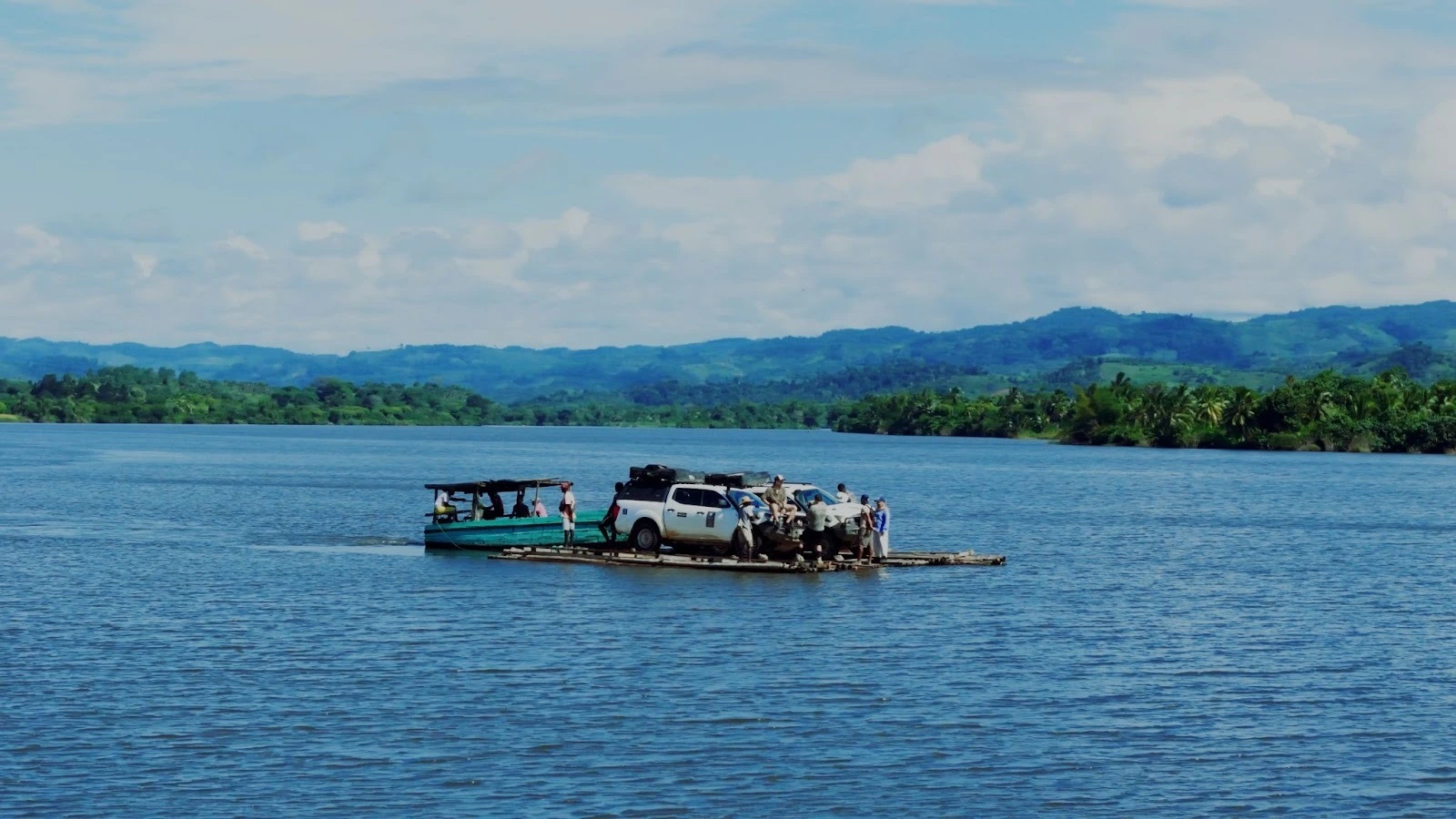 n off-road pickup truck loaded with overland gear being transported across a wide river on a makeshift wooden raft pushed by a small boat, with lush green hills and a blue sky in the background along Route Nationale 5 in Madagascar.