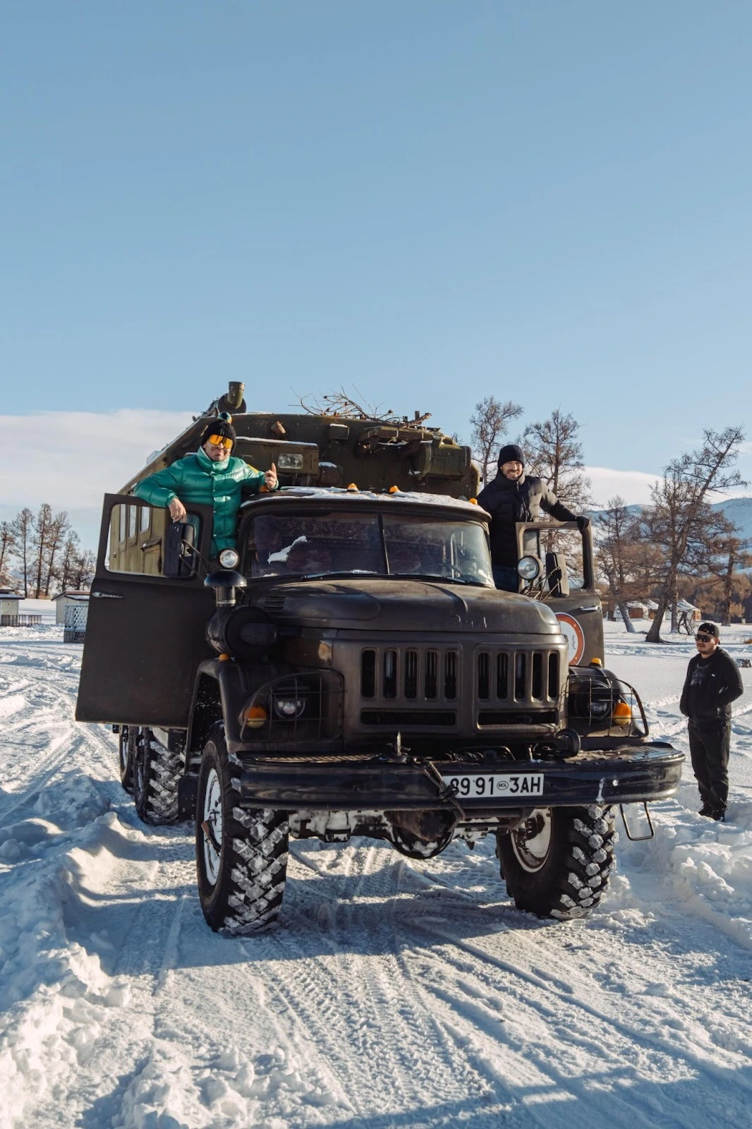 A rugged, vintage 6x6 military truck parked on a deep, snow-covered track under a clear blue sky, with three people standing on and next to the vehicle during a winter expedition.